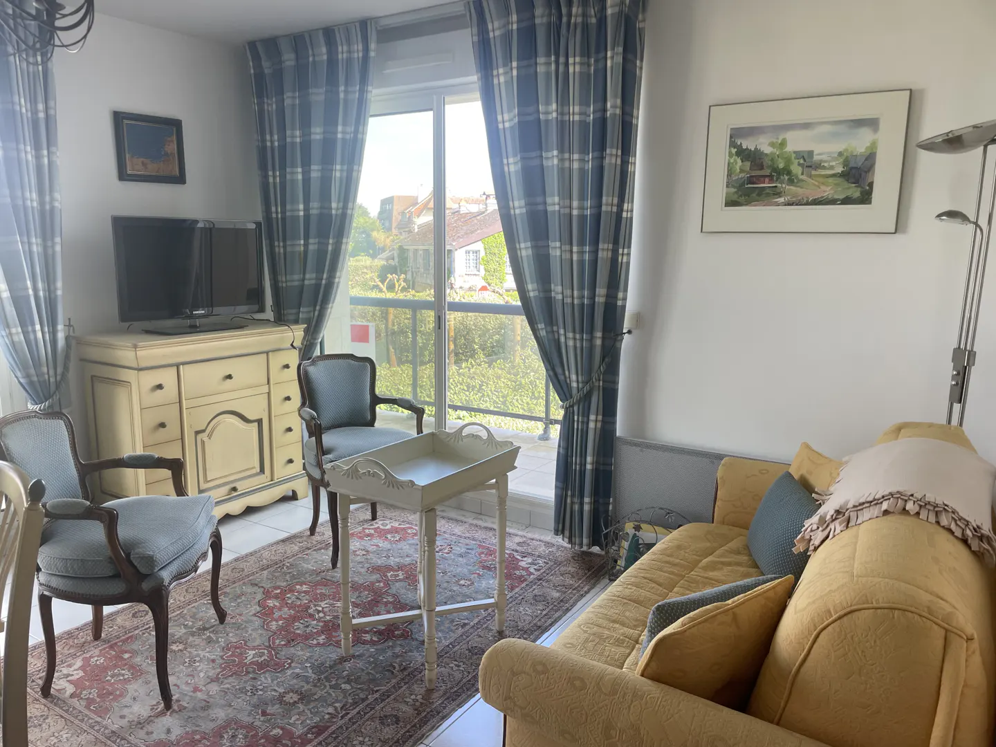 Living room with blue plaid curtains, a yellow sofa, and two blue chairs on a red patterned rug. A TV sits on a cream-colored dresser.