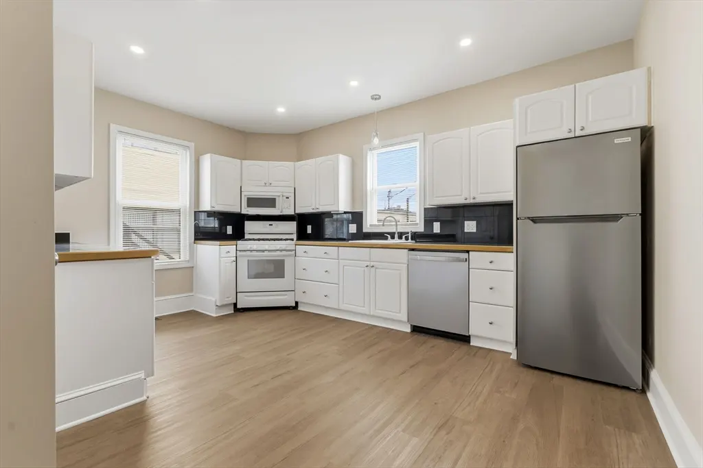 Bright kitchen with white cabinets, stainless steel appliances, and light wood-look flooring. Black backsplash and light tan walls.