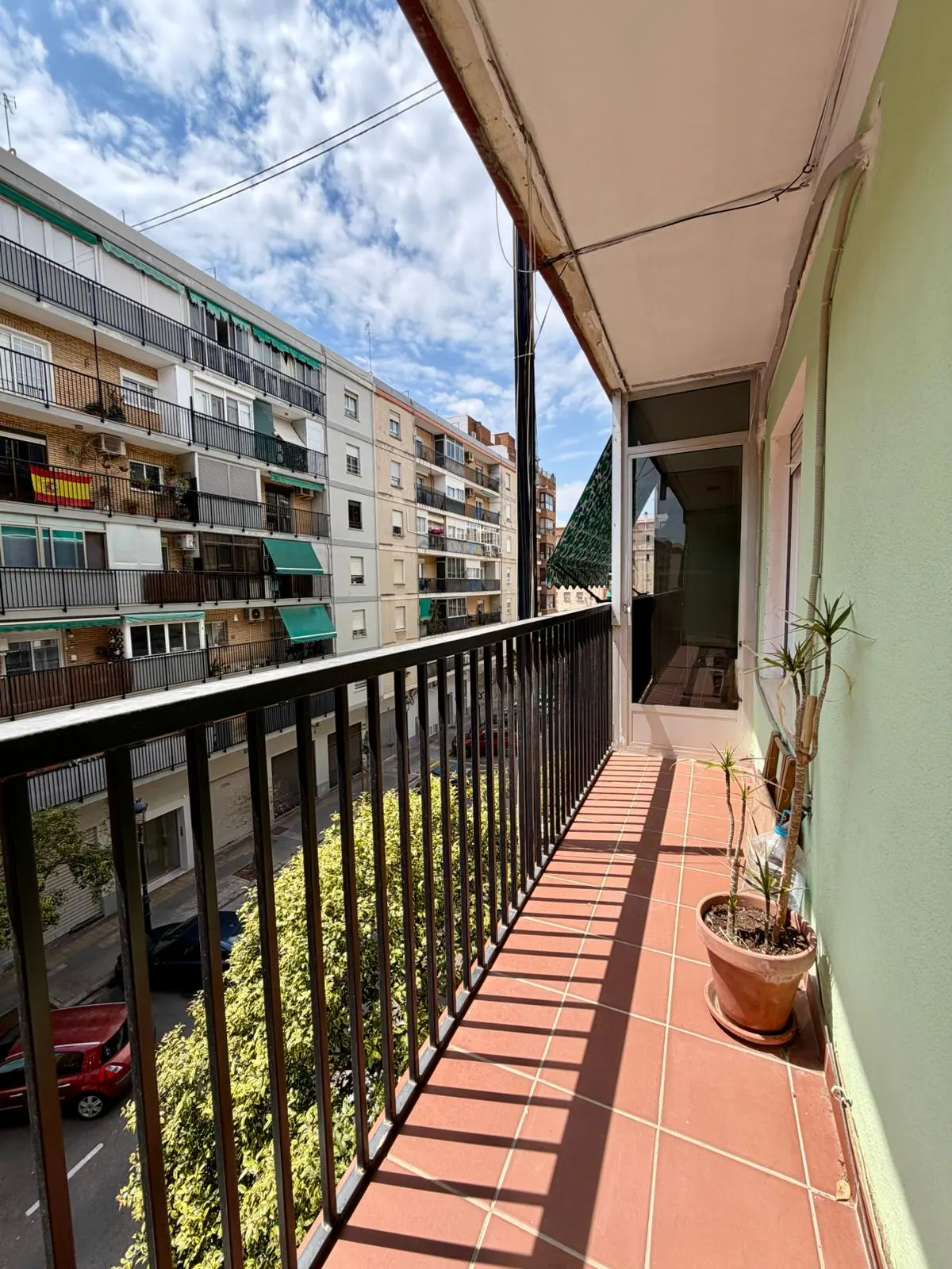 A sunny balcony with terracotta tiles, a black railing, and a potted plant, overlooking a city street with apartment buildings.