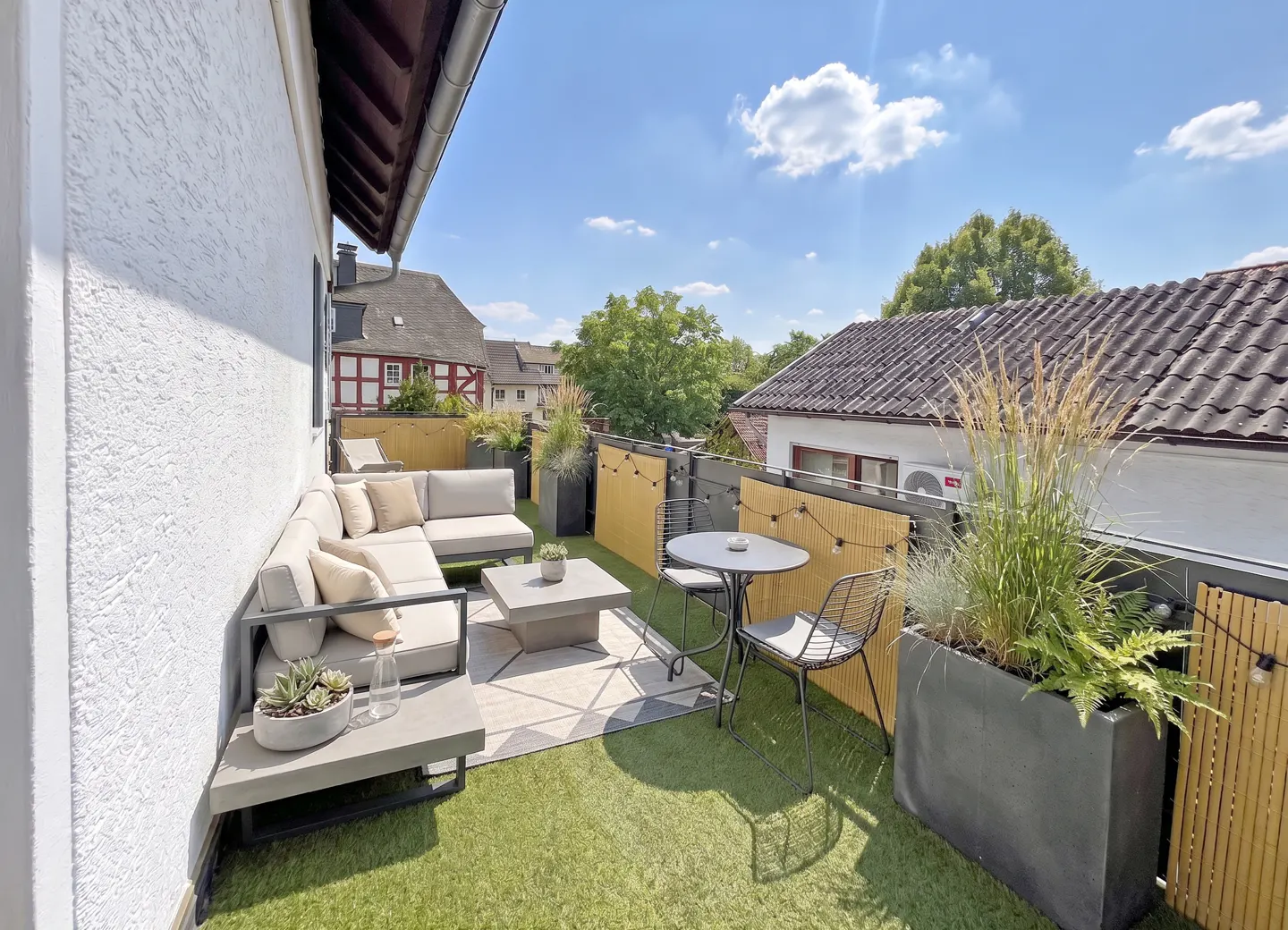 Outdoor patio with a gray sectional sofa, table, chairs, and plants on artificial grass, with a blue sky in the background.