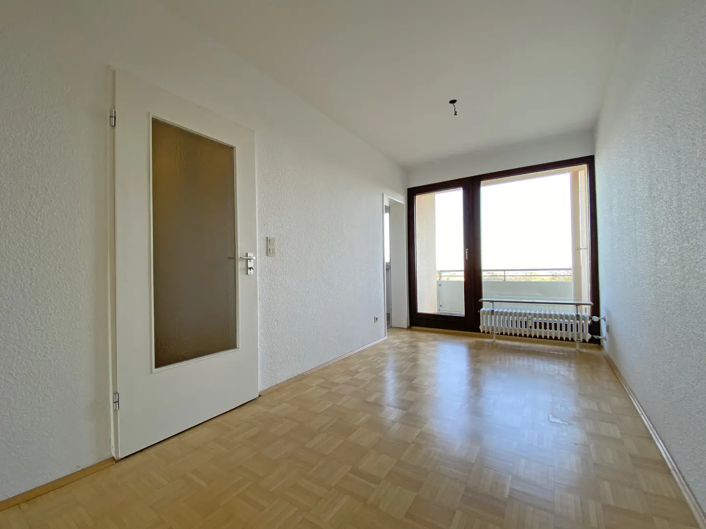 Bright, empty room with parquet floors, white walls, and a door to a balcony. A white door with a glass panel is on the left.