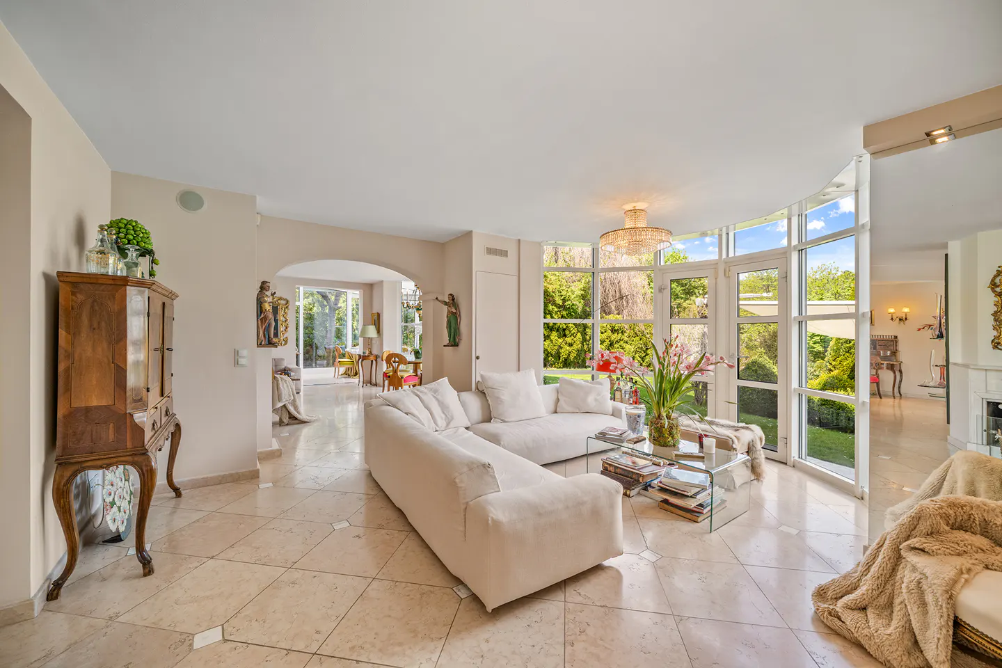 Bright living room with marble floors, a white sectional sofa, and floor-to-ceiling windows overlooking a green garden. A glass coffee table sits in front of the sofa.