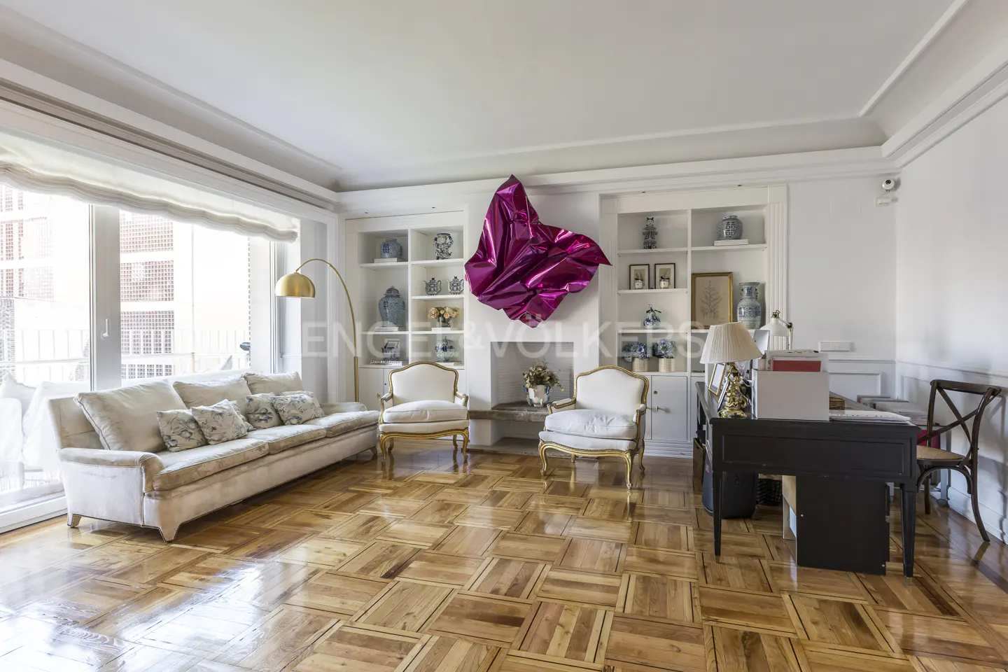 Bright living room with parquet floors, a cream sofa, two chairs, and a desk. A large, shiny pink sculpture hangs above the fireplace.