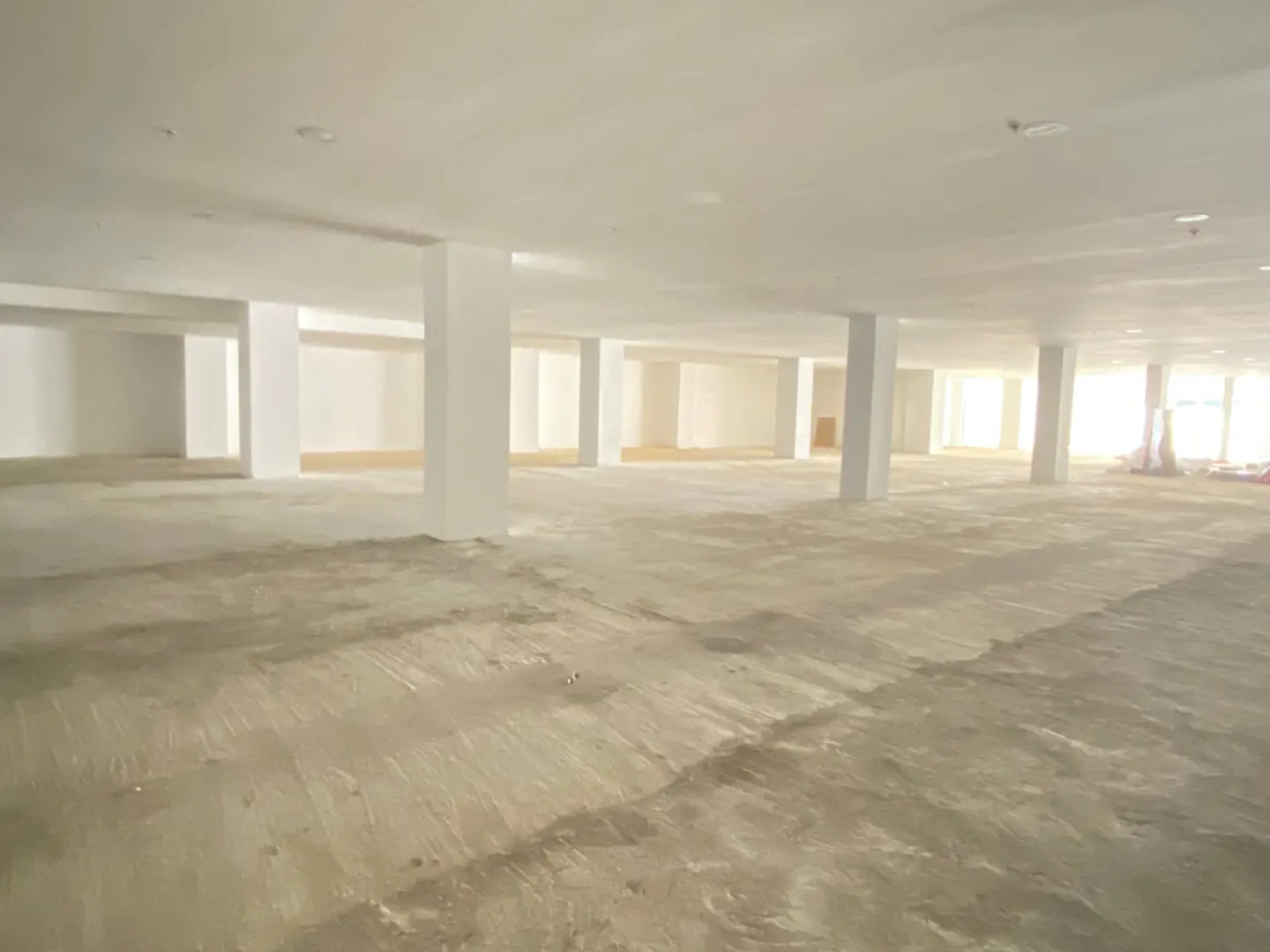A large, empty commercial space with white walls, concrete floors, and support pillars under bright interior lighting.
