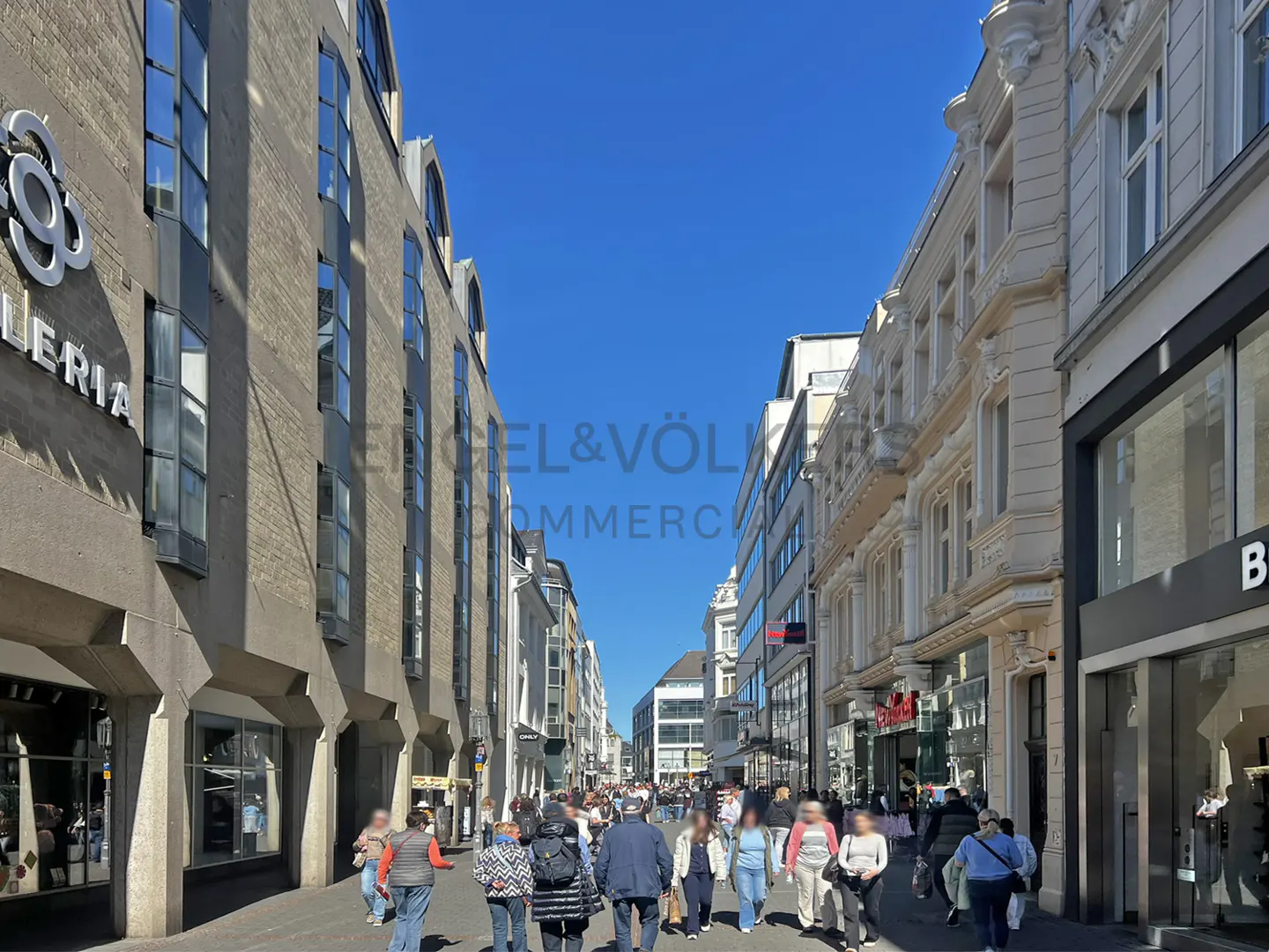 Street view of a busy pedestrian zone with shops and buildings on both sides under a clear blue sky.