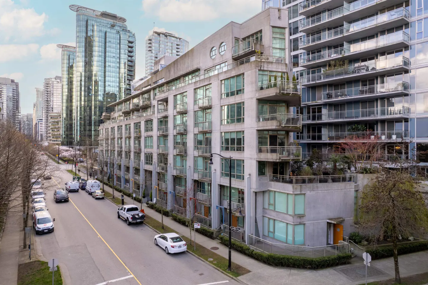 Exterior view of a modern, multi-story apartment building with large windows and balconies, street with parked cars, and skyscrapers in the background.