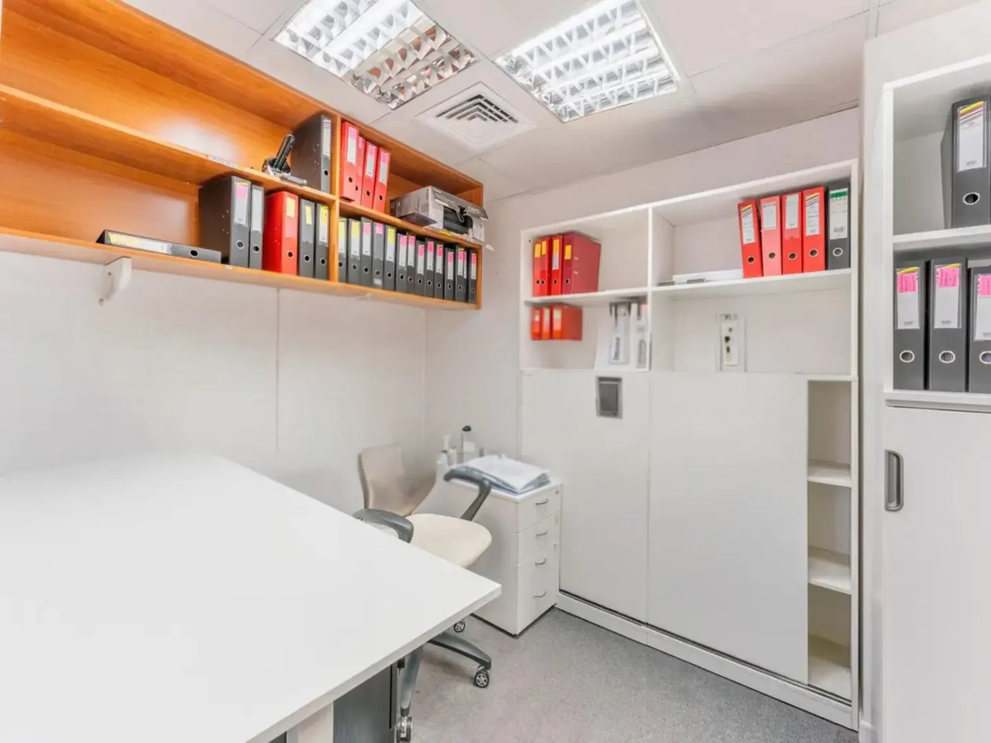 Office space with white walls, a large white desk, and a rolling chair. Shelves hold red and black binders.