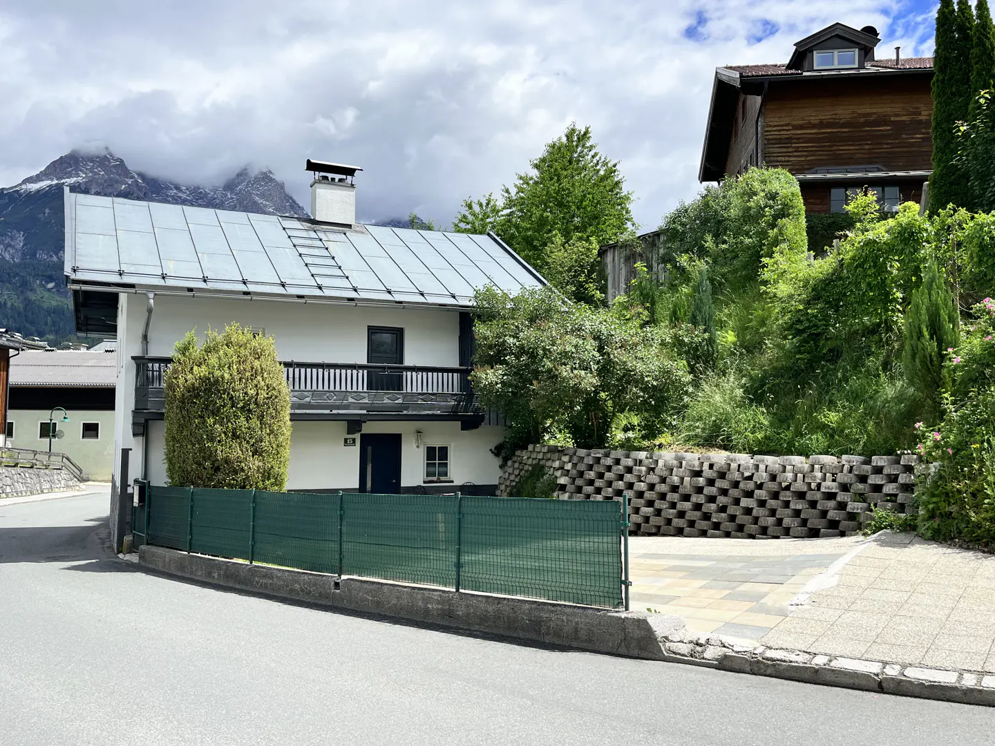White house with a metal roof, balcony, and green fence, set against a backdrop of mountains and lush greenery.