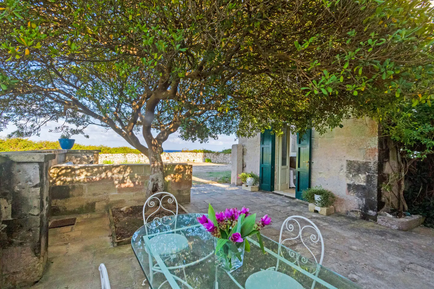 Outdoor patio with glass table, white chairs, and purple flowers under a large tree. Stone building with green shutters in the background.