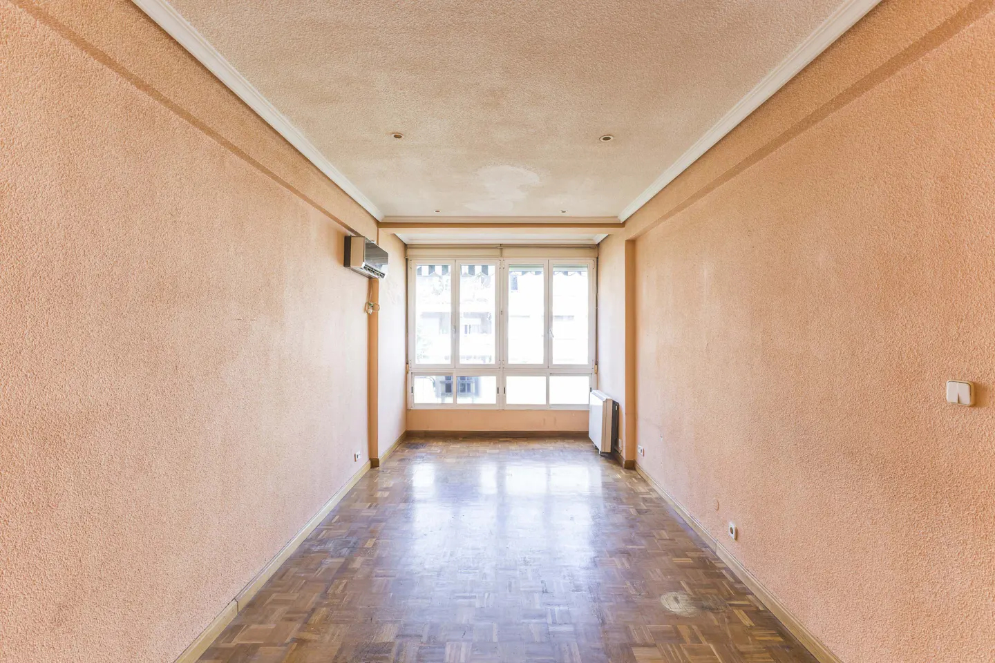Empty room with peach walls, parquet floor, and a large window. An air conditioner is mounted high on the left wall.