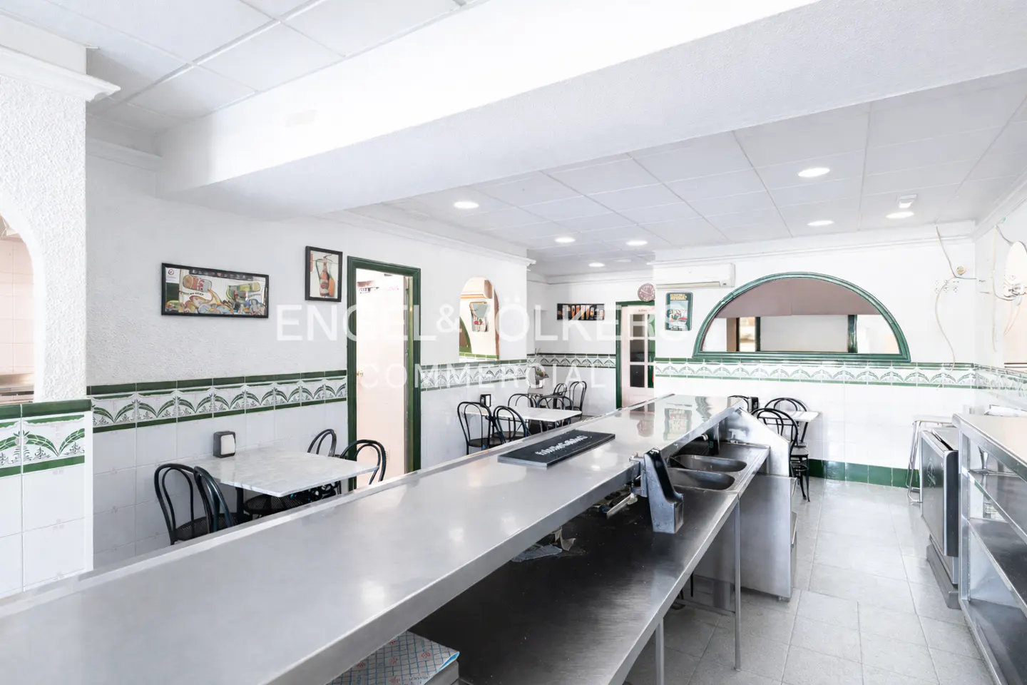 Interior view of a restaurant with a long stainless steel bar, tables, and white walls with green tile accents.