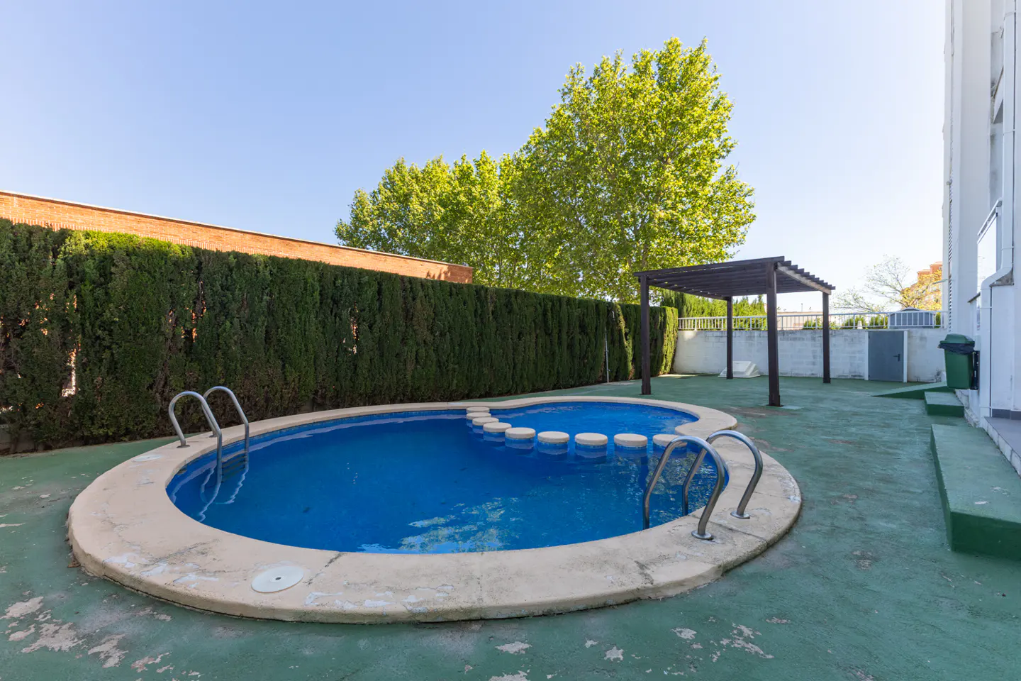 Backyard view of a kidney-shaped pool with blue water, surrounded by green turf, a hedge, and a pergola.