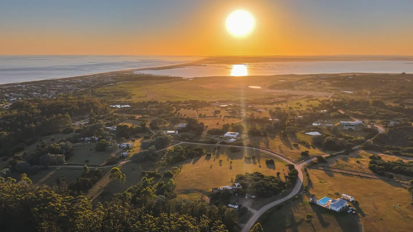 Aerial view of a coastal community at sunset, featuring homes, trees, and a winding road. The sun reflects on the water.