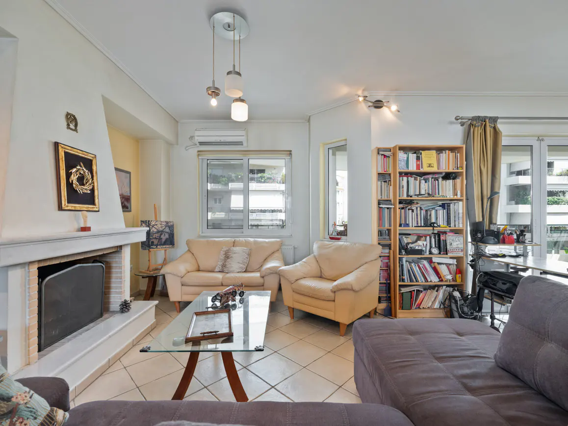 Living room with beige sofas, a glass coffee table, and a fireplace with a black screen. A bookcase stands near a window.
