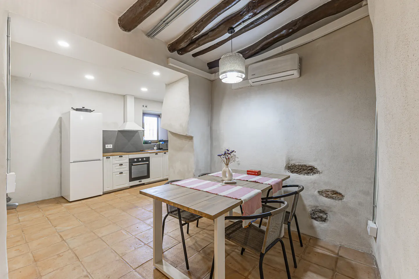 Rustic kitchen and dining area with terracotta tile floor, white cabinets, wooden beams, and a table set for two.