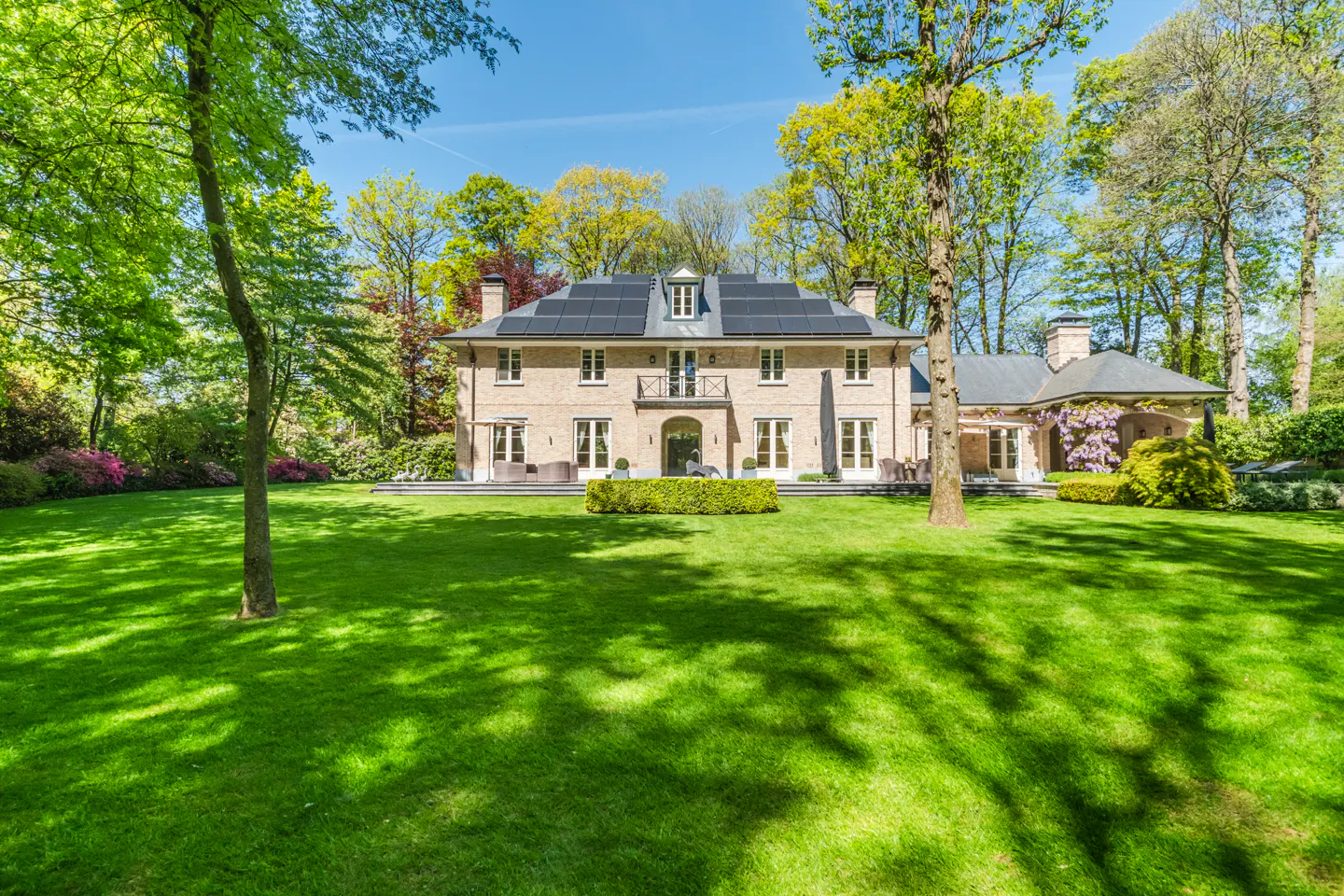 Beige brick house with solar panels, surrounded by green trees and lawn under a blue sky.