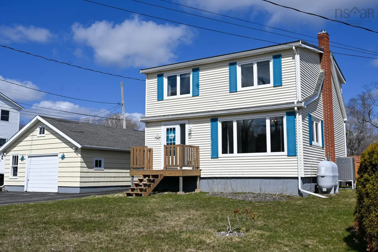 Two-story house with cream siding and blue shutters, a brick chimney, and a detached garage on a sunny day.