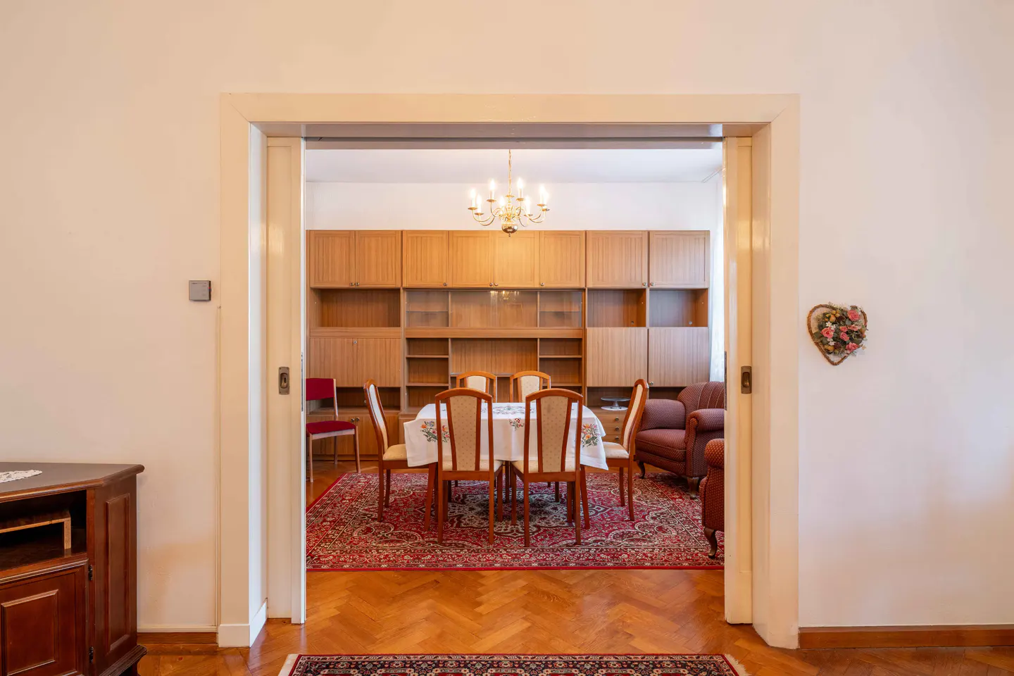 View through doorway into dining room with table, chairs, and built-in wooden cabinets. Red patterned rug on wood floor.