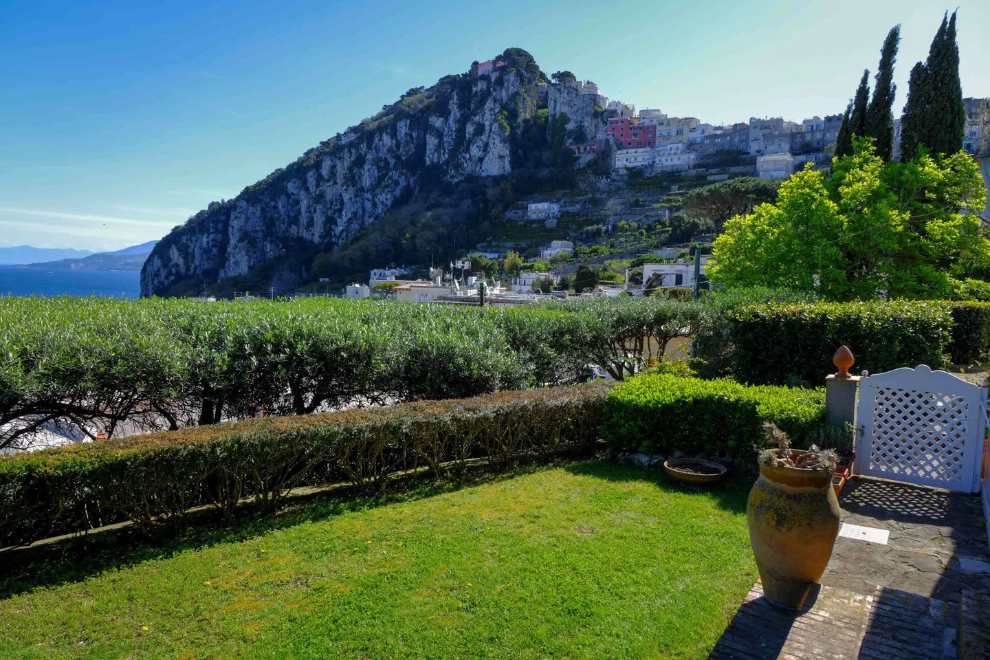 Scenic view of a lush green lawn with a mountain backdrop and a town on the hillside. A large pot sits near a white gate.