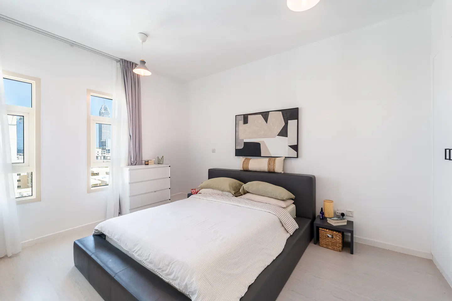 Bright bedroom with a gray platform bed, white bedding, and green pillows. Windows show a city view. Abstract art hangs above the bed.
