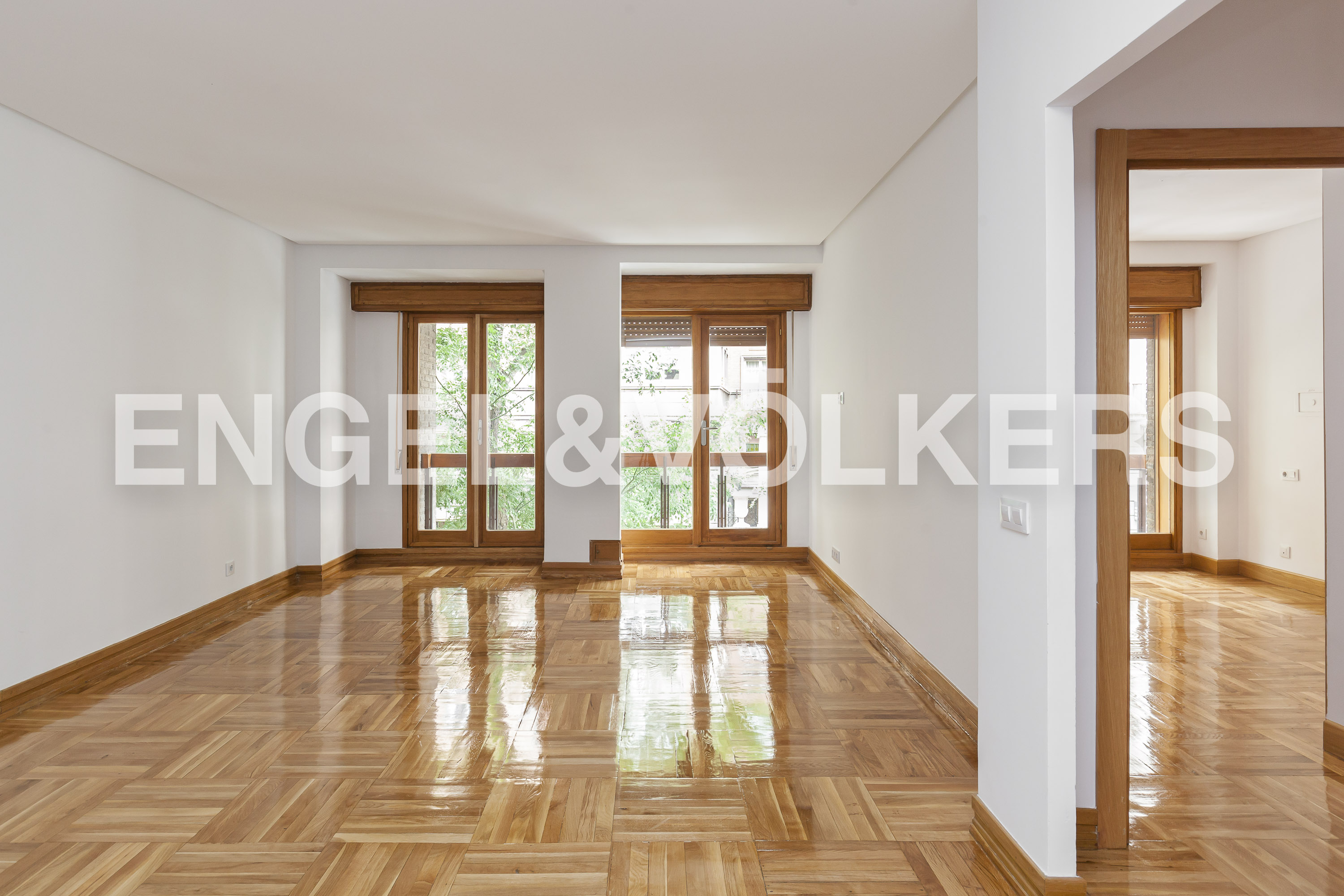 Bright, empty room with white walls, wood floors, and two windows with wood frames. A doorway leads to another room with similar features.