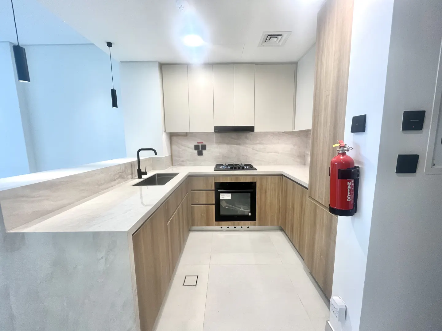 A modern kitchen with light wood cabinets, white countertops, and a black faucet. A red fire extinguisher is mounted on the wall.