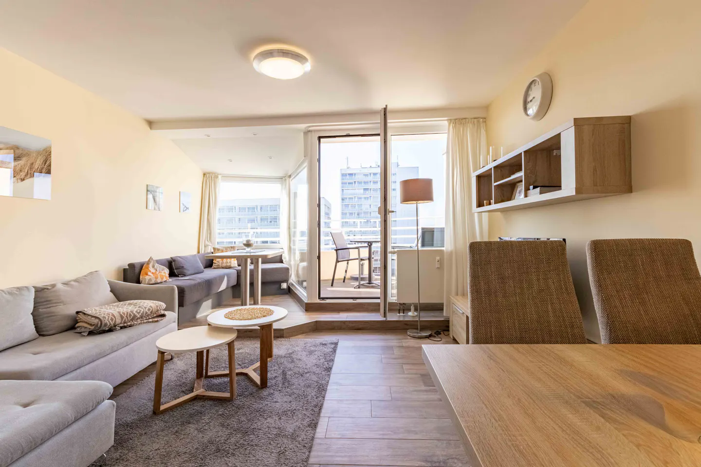 Living room with gray sofas, wood floors, and a balcony with city views. A dining table and chairs are in the foreground.