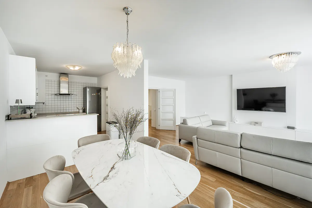 Bright, open-concept apartment with white walls, wood floors, and modern furnishings. A marble dining table sits near a gray sofa and a kitchen area.