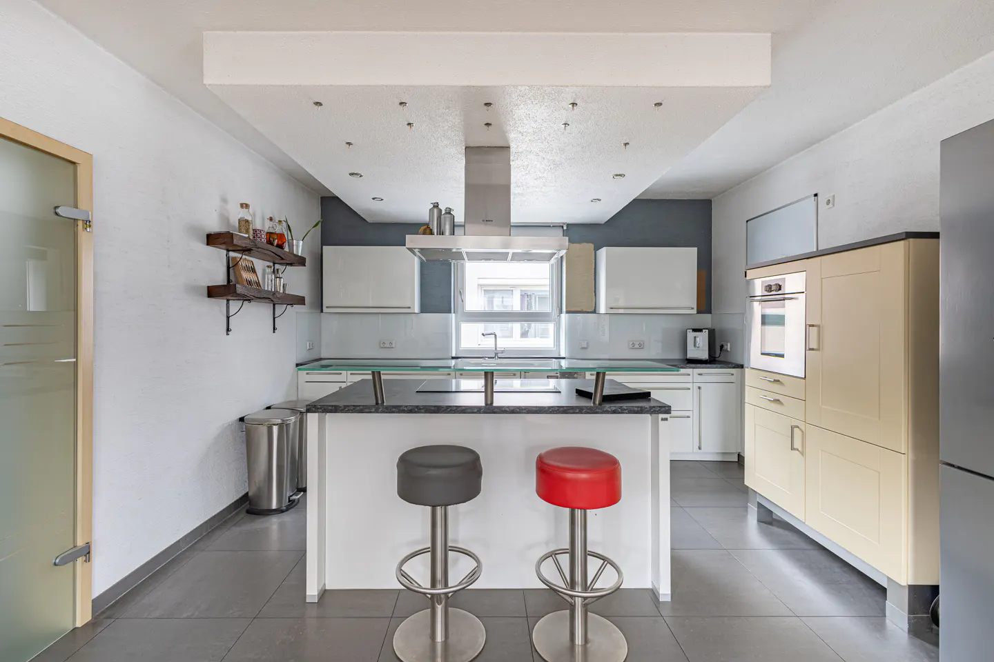 A modern kitchen with white cabinets, a gray accent wall, and a central island with two stools, one red and one gray.