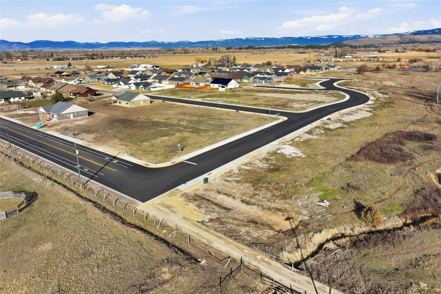 Aerial view of a new housing development with black paved roads, houses, and a mountain backdrop under a blue sky.