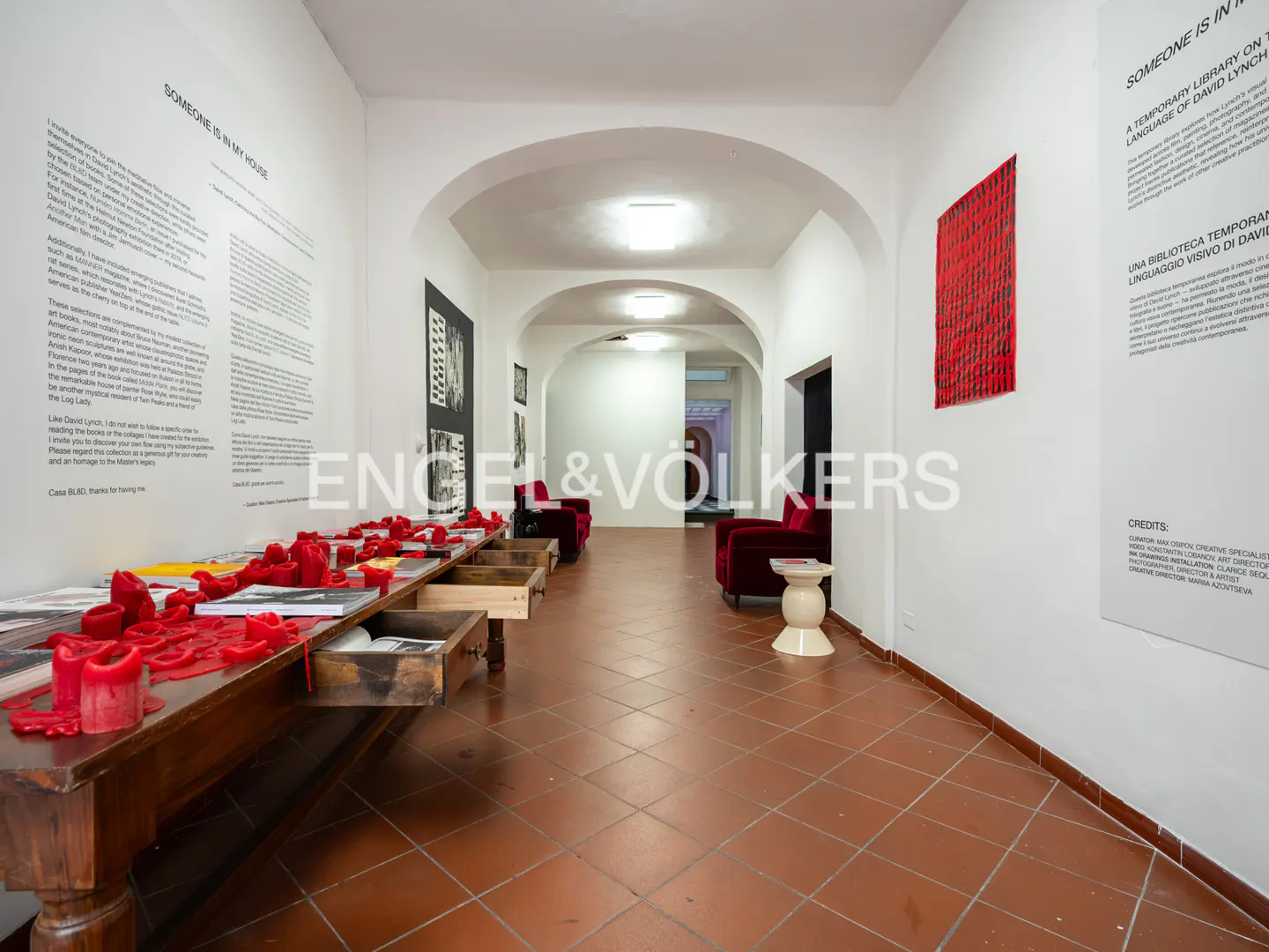 A gallery hallway with a long wooden table covered in red wax, red armchairs, and informational text on white walls.