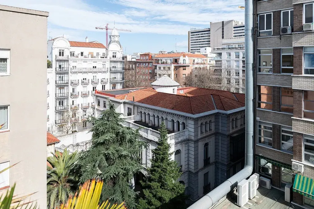 Cityscape view with white and brown buildings, green trees, and a blue sky with clouds. A construction crane is visible in the distance.