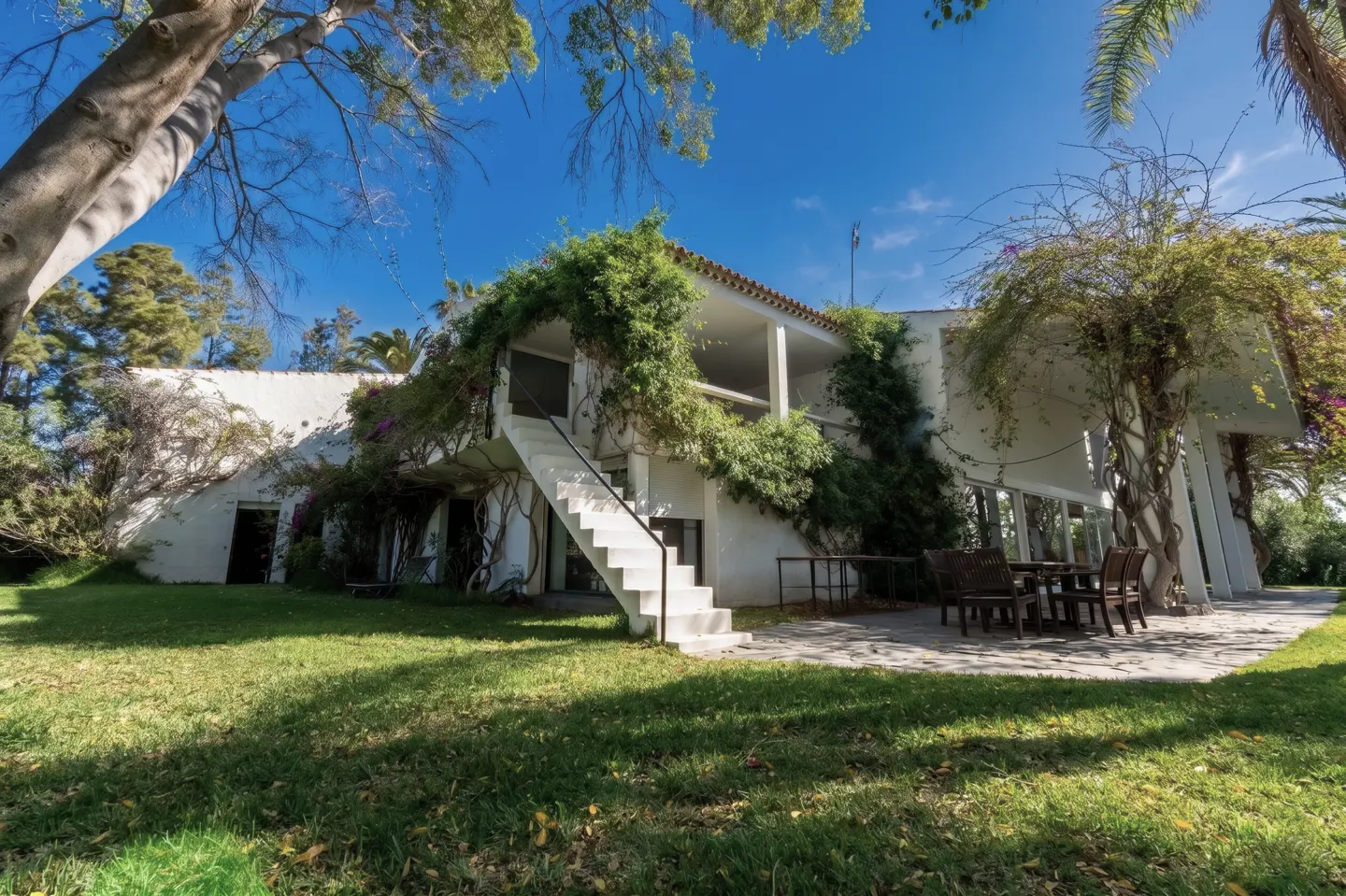 Exterior of a white two-story house with a staircase, patio, and green lawn under a blue sky.