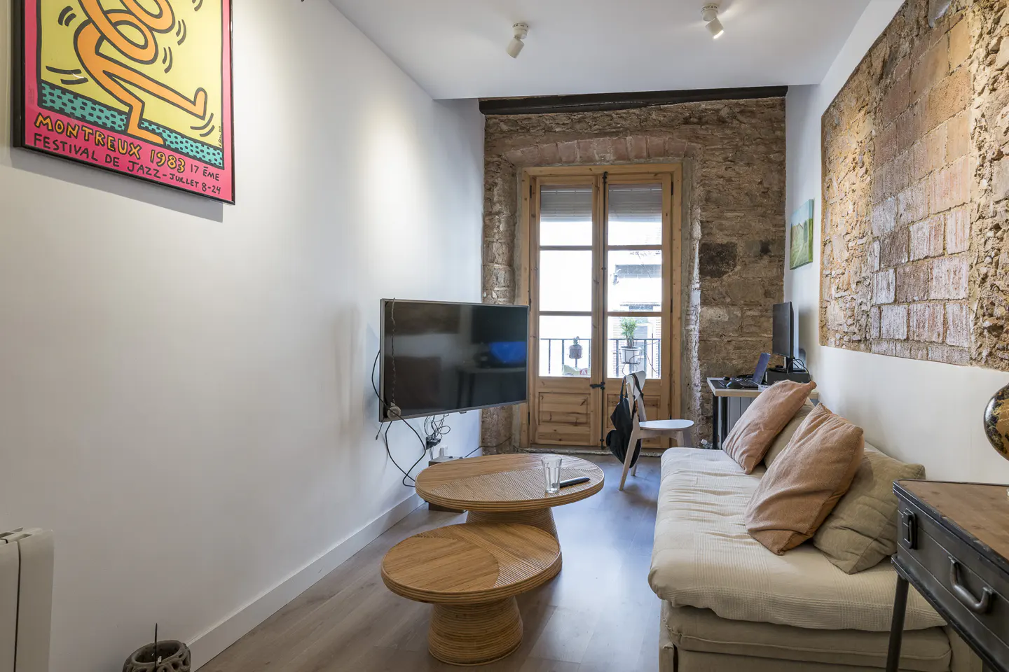 Cozy living room with white walls, exposed brick, and a wooden door to a balcony. A Keith Haring print hangs above a TV and wooden tables.