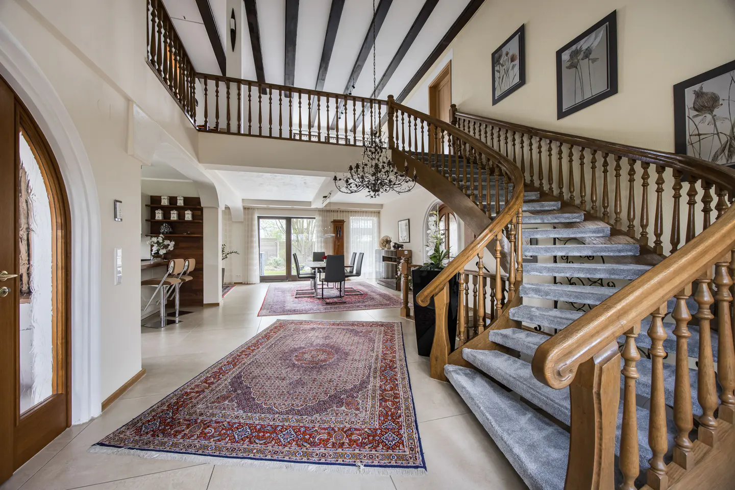 Grand foyer with a curved wood staircase, gray carpet, and a large patterned rug. A dining area is visible in the background.