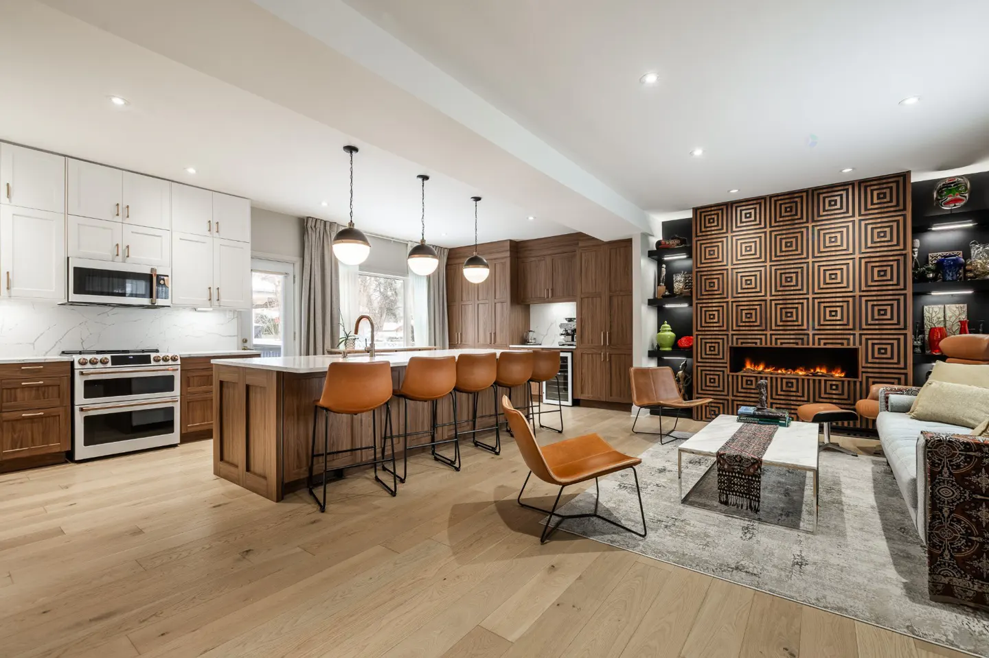 Open-concept kitchen and living room with wood floors. Kitchen has white cabinets, island with brown stools. Living room has a fireplace and sofa.