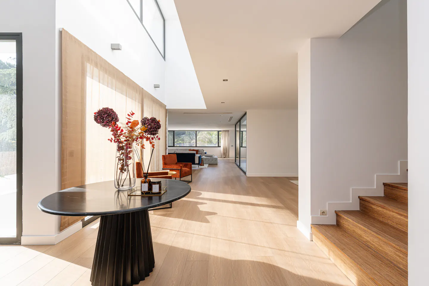 Bright, modern home foyer with wood floors, white walls, and a black accent table with flowers. Living room visible in background.