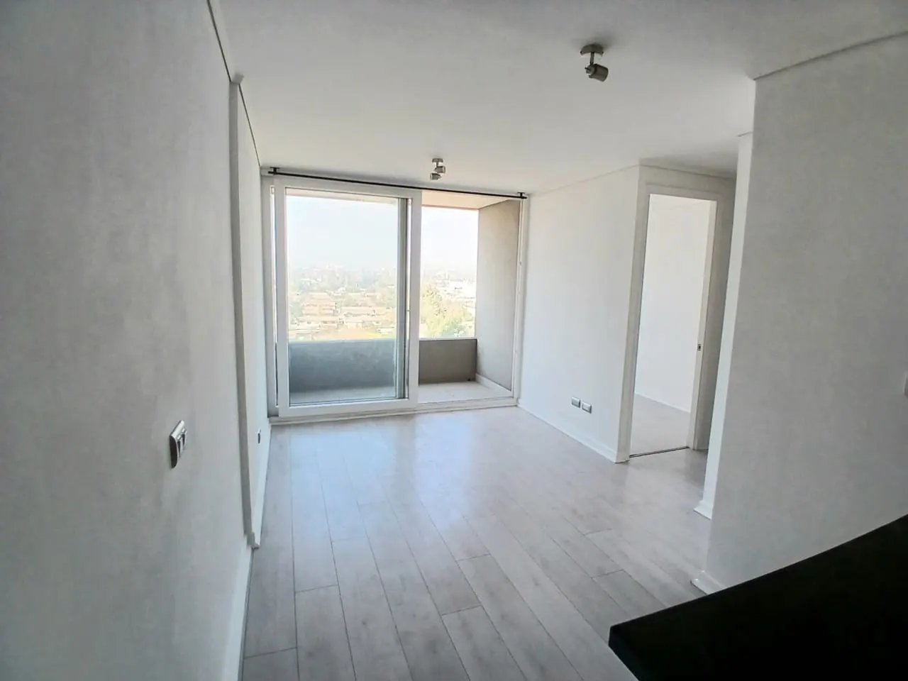 Bright, empty apartment with white walls, light wood floors, and a sliding glass door to a balcony overlooking a cityscape.