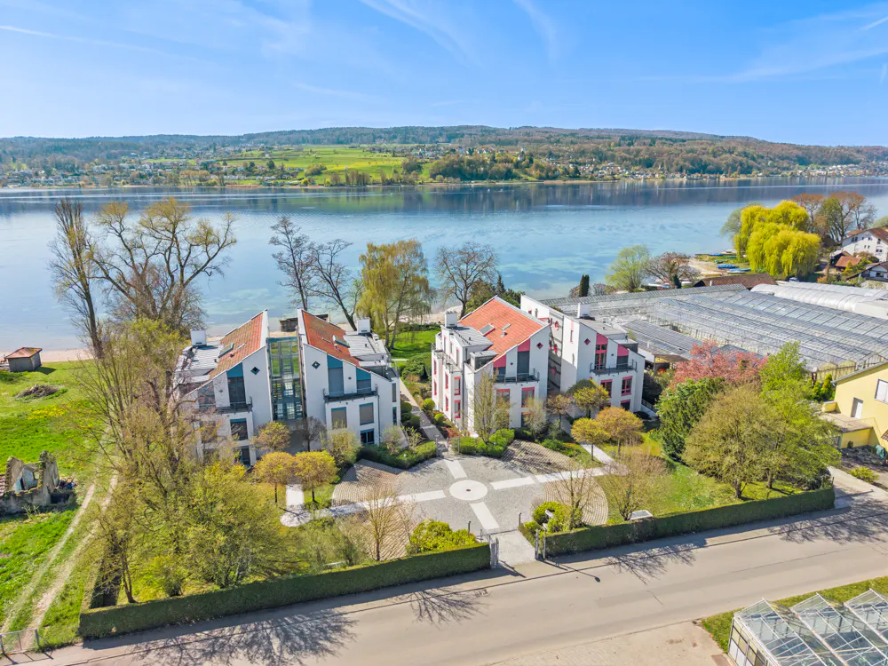Aerial view of two modern white buildings with red roofs near a lake, surrounded by green trees and lawns.