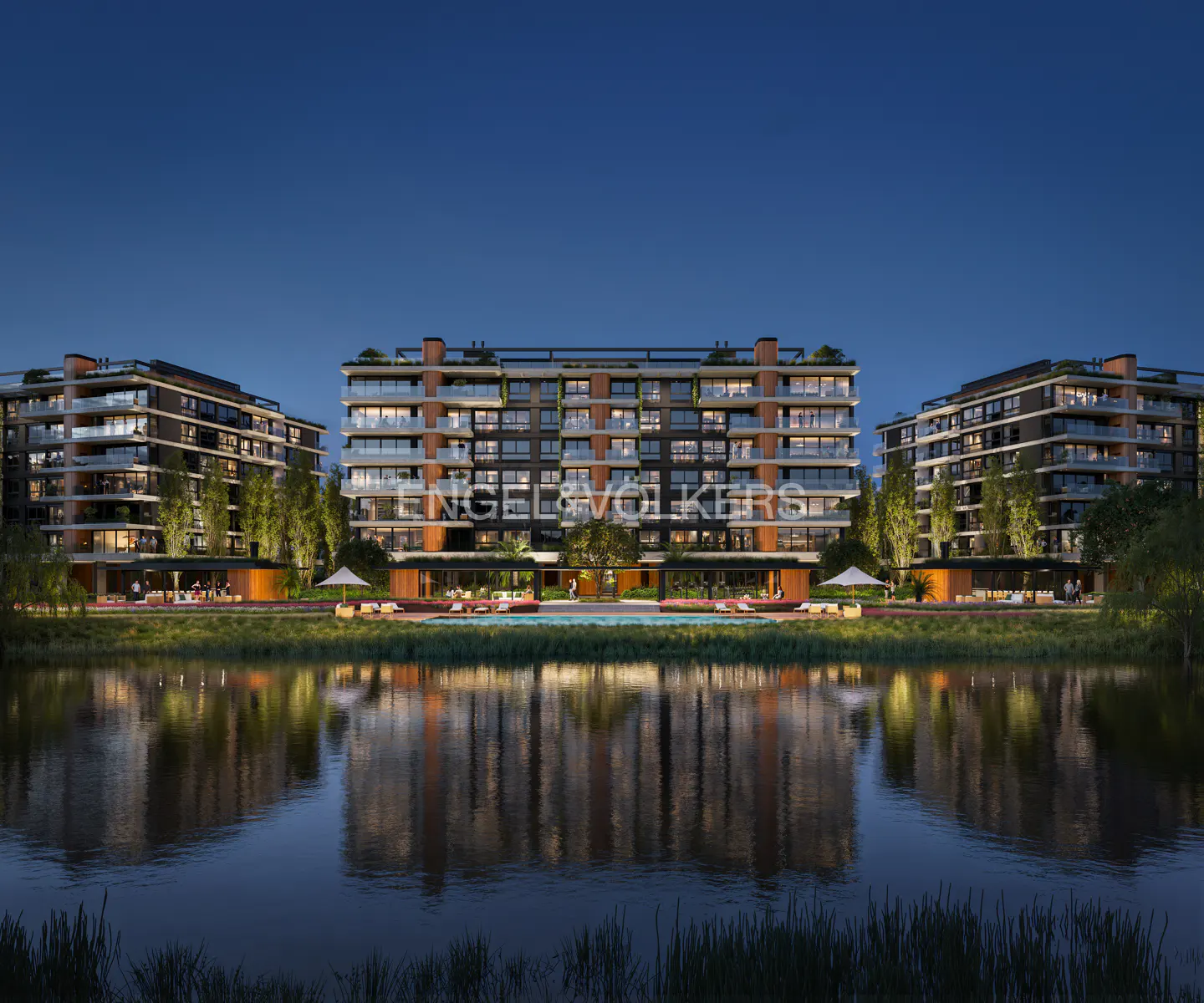 Three modern apartment buildings with balconies face a lake at dusk, their reflections visible in the water.
