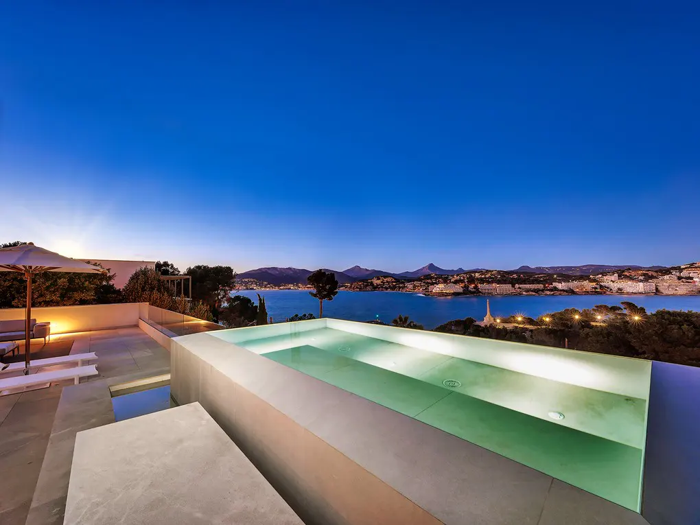 Illuminated infinity pool on a terrace at dusk, overlooking a bay with distant mountains and a coastal town.