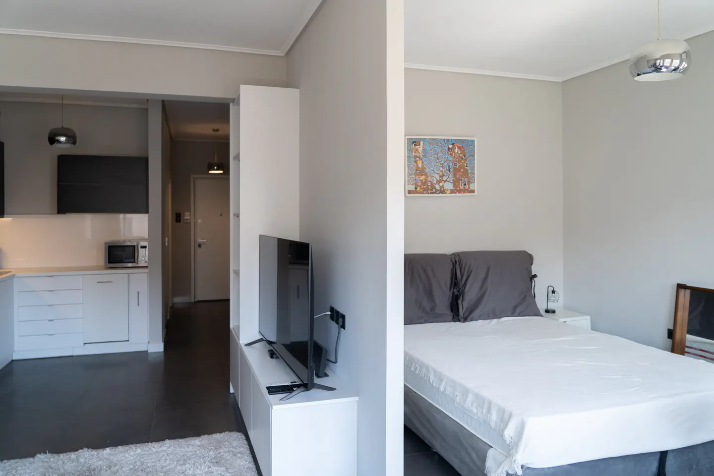 Modern studio apartment with a white kitchen, a TV on a white console, and a bedroom area with a bed and wall art.