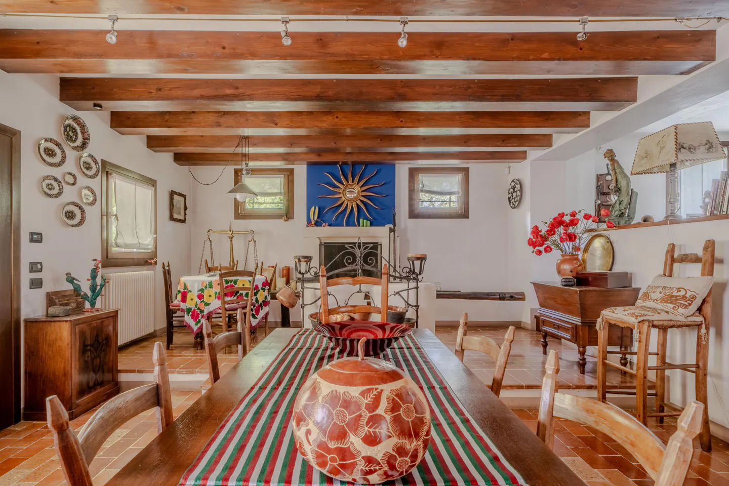 Rustic dining room with a long wooden table, chairs, and a striped runner. Beamed ceiling, fireplace with sunburst art, and terracotta tile floor.