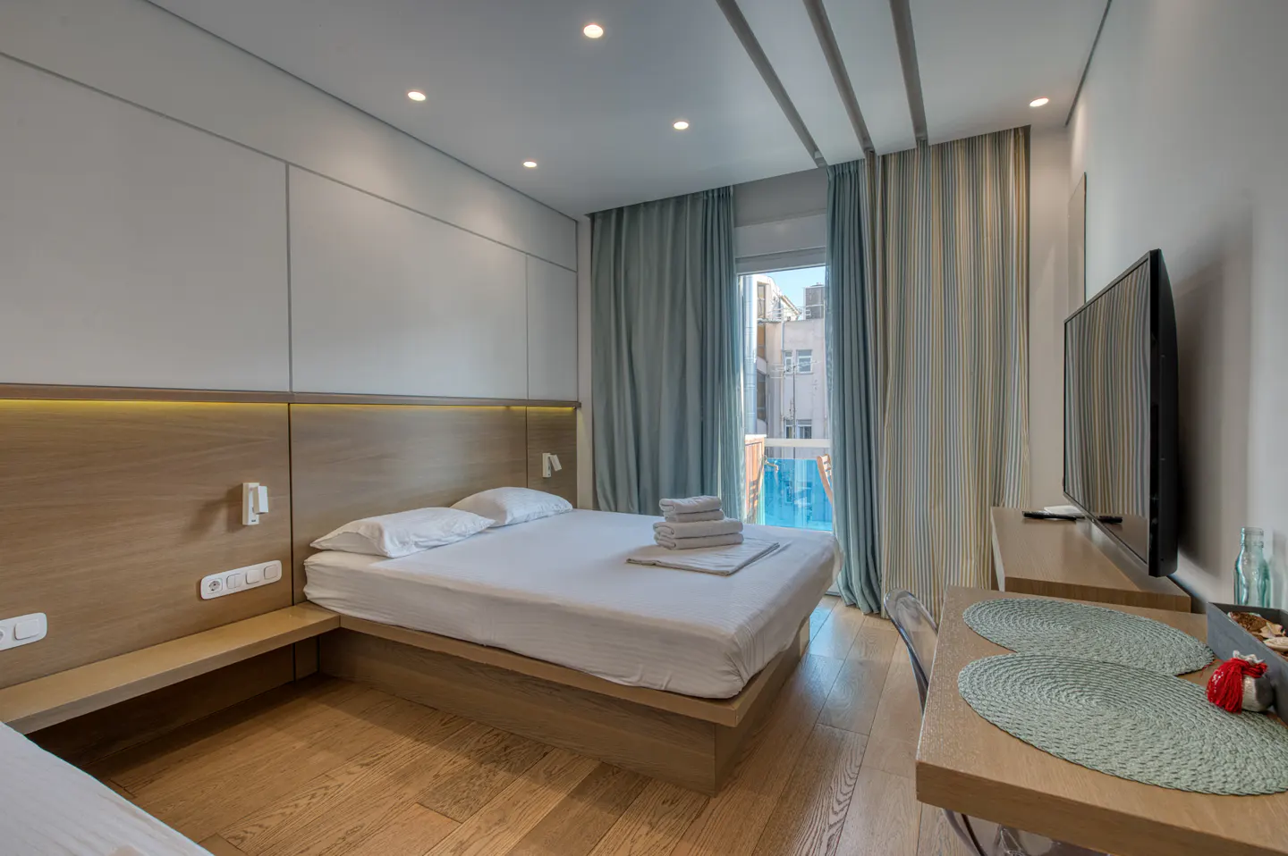Modern bedroom with a queen bed, white linens, and a wooden headboard. A TV sits on a wooden dresser, and blue curtains frame a window with a pool view.