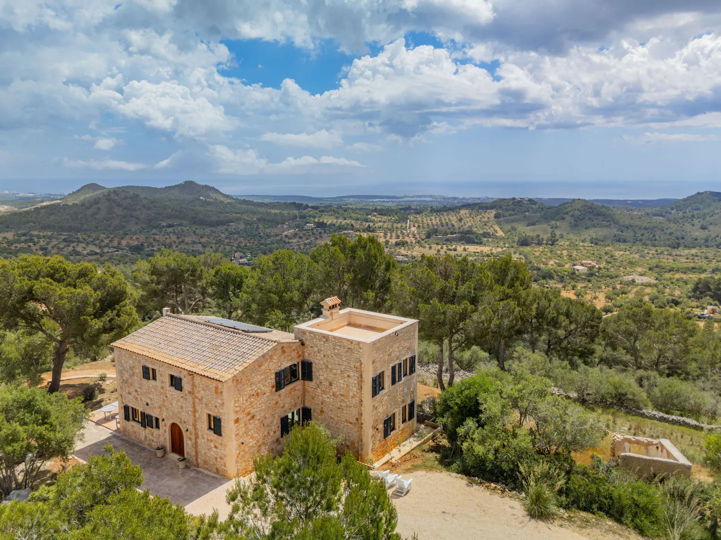 Aerial view of a stone house with a tile roof, surrounded by green trees and rolling hills under a cloudy sky.