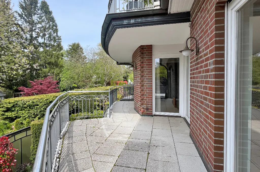 Curved balcony with gray stone tiles, a metal railing, and a brick wall. Green trees and bushes are visible in the background.