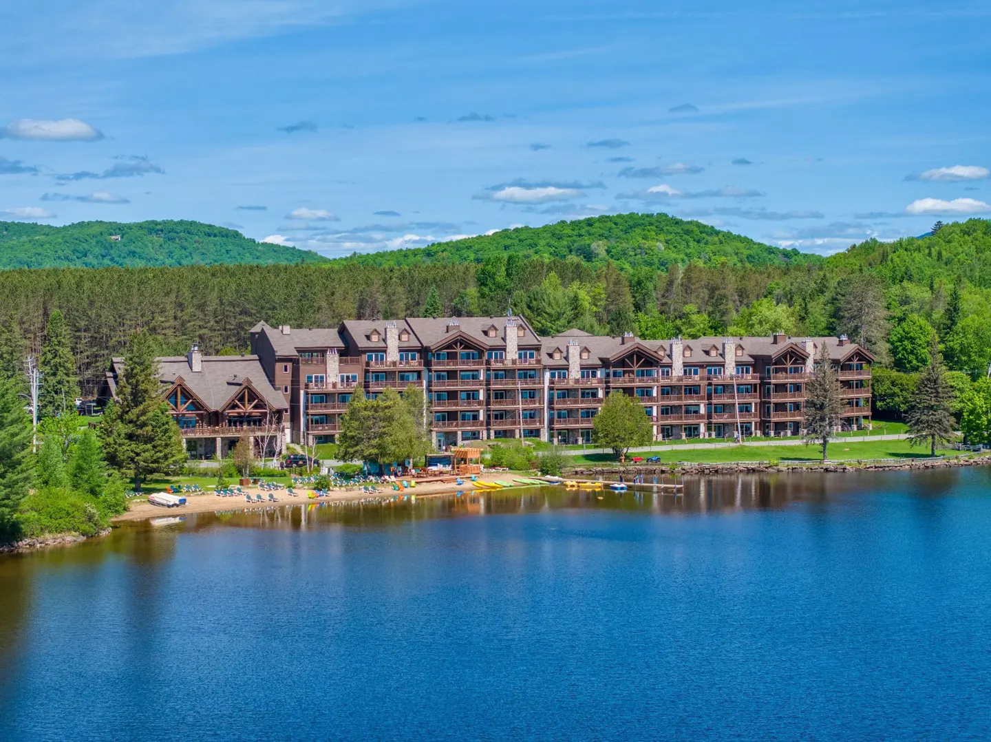 Scenic view of a lakeside resort with a sandy beach, kayaks, and lush green mountains in the background under a blue sky.