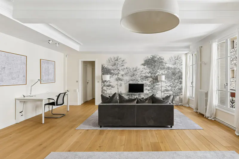 Bright living room with wood floors, white walls, and a gray sofa facing a black and white forest mural. A white desk and chair are on the left.