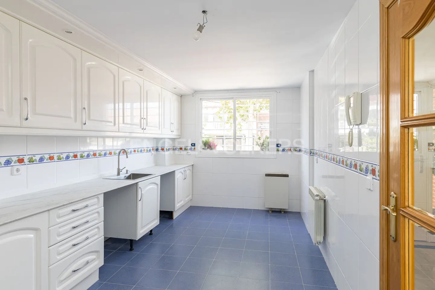 Bright kitchen with white cabinets, blue tile floor, and white tiled walls with a fruit-themed border. A window lets in natural light.