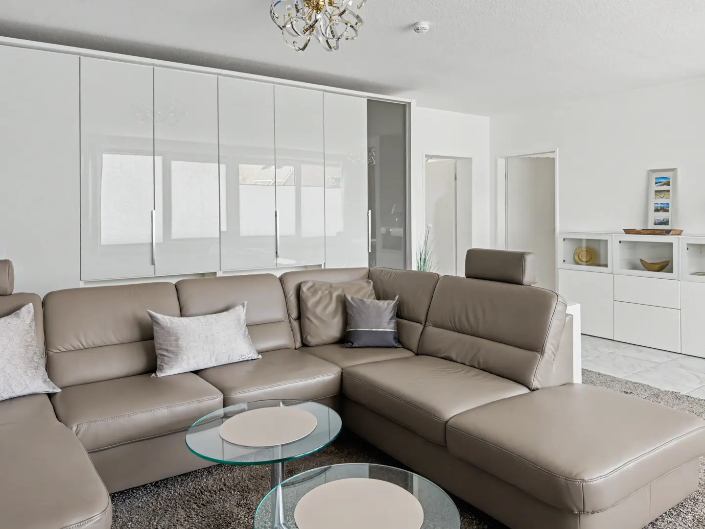 Living room with a taupe leather sectional sofa, glass coffee tables, and white cabinets. A chandelier hangs from the ceiling.